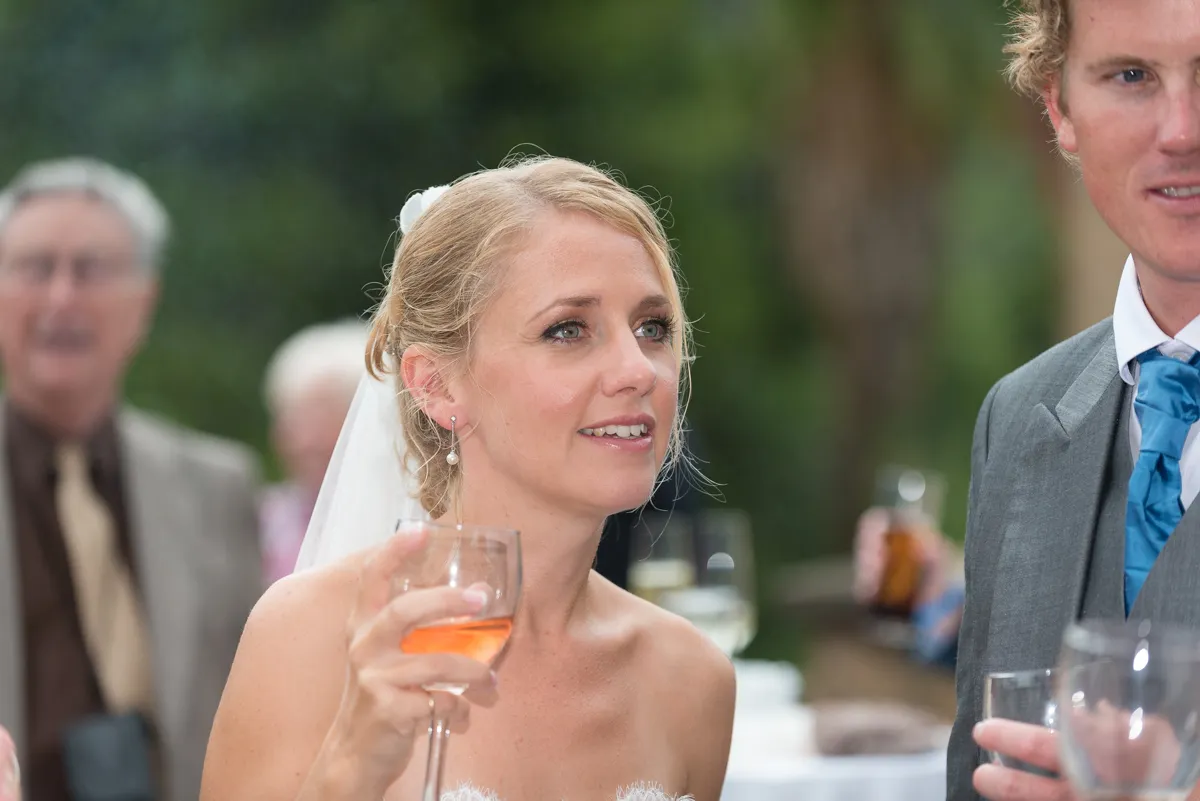 Bride in a strapless white dress holding a glass of rosé wine and looking at the groom during an outdoor wedding reception.