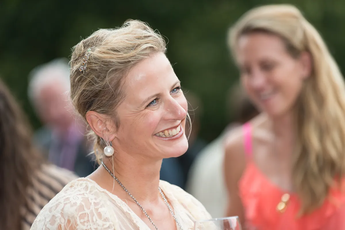 Smiling woman with blonde hair and pearl earrings wearing a lace top, holding a glass with blurred people in the background.