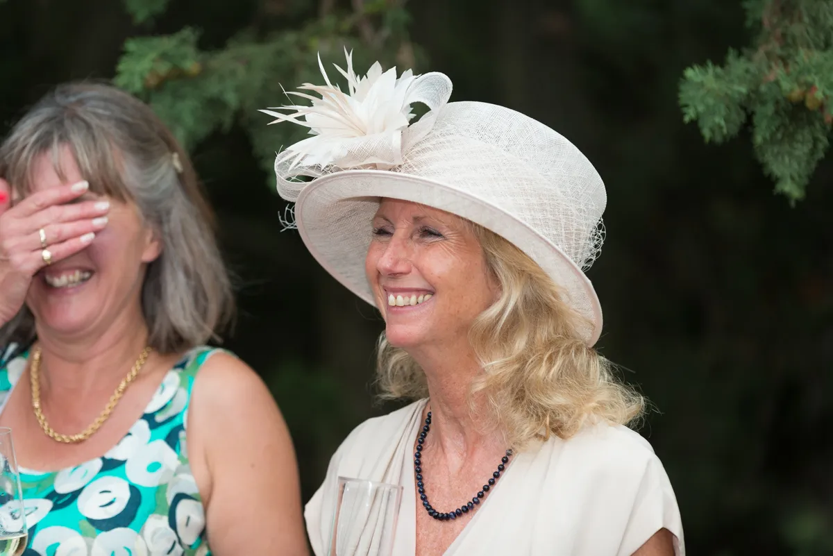 Two women smiling, one wearing a white decorative hat and holding a glass of champagne, the other laughing with her hand covering part of her face.