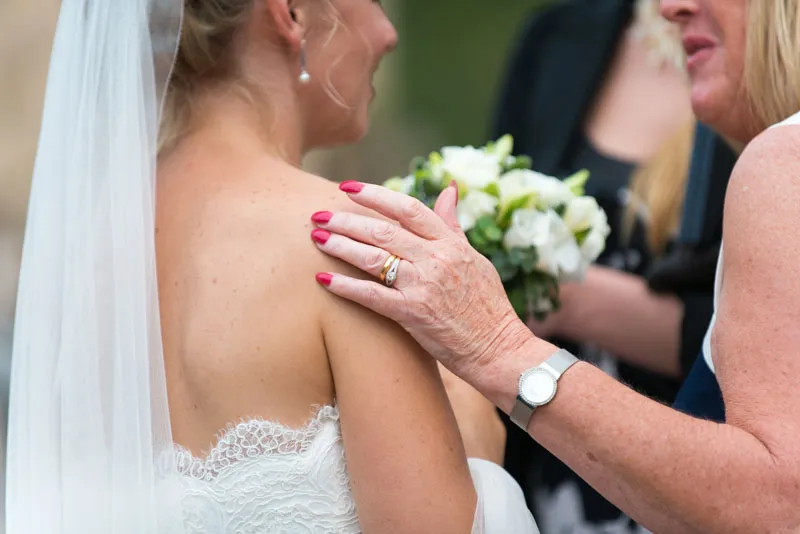 Close-up of a bride in a strapless lace wedding dress with a veil, gently touched on the shoulder by an older woman wearing a watch and wedding rings.