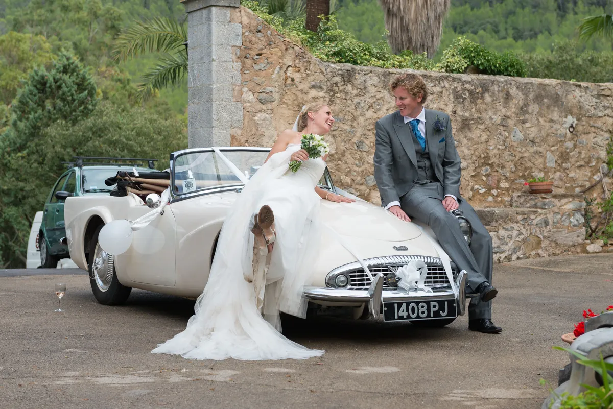Bride in a white wedding dress holding a bouquet and groom in a gray suit sitting on the hood of a decorated vintage white car, smiling at each other.