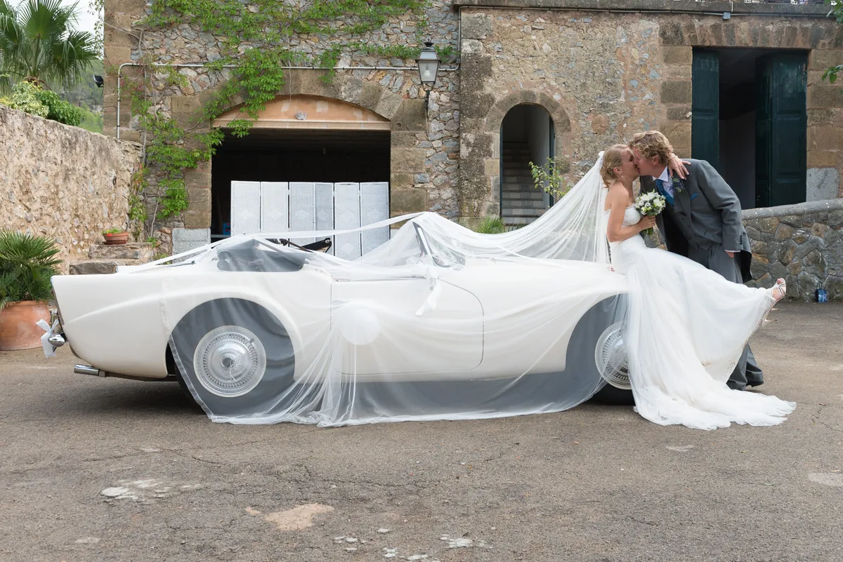 Bride and groom kissing beside a white vintage convertible car draped with bride's long veil in front of a stone building.