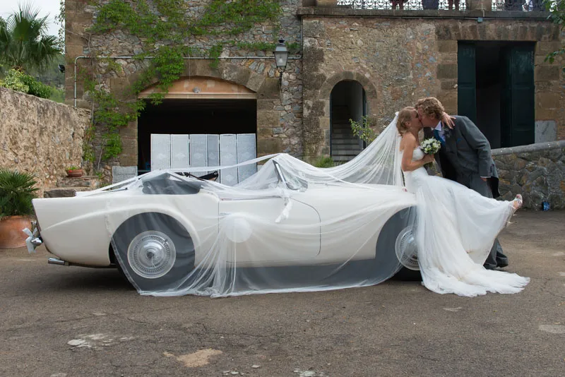 Bride and groom kissing beside a vintage white convertible car draped with the bride's long veil.