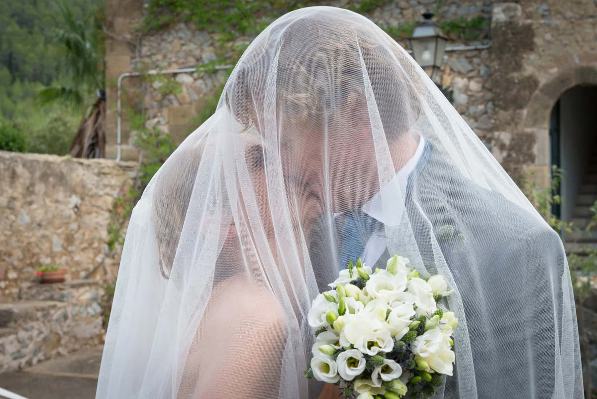 Bride and groom embracing closely under a white veil, with the groom holding a bouquet of white flowers.