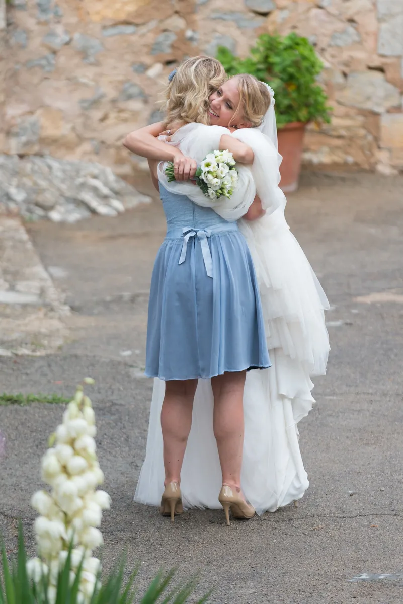 Bride in a white wedding dress hugging a woman in a blue dress while holding a bouquet of white flowers.