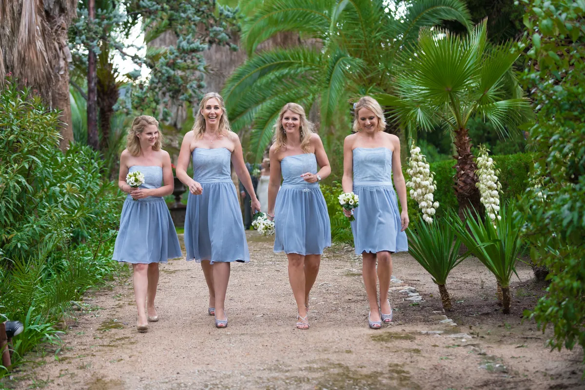 Four women in matching light blue strapless dresses walking down a garden path holding small white flower bouquets.