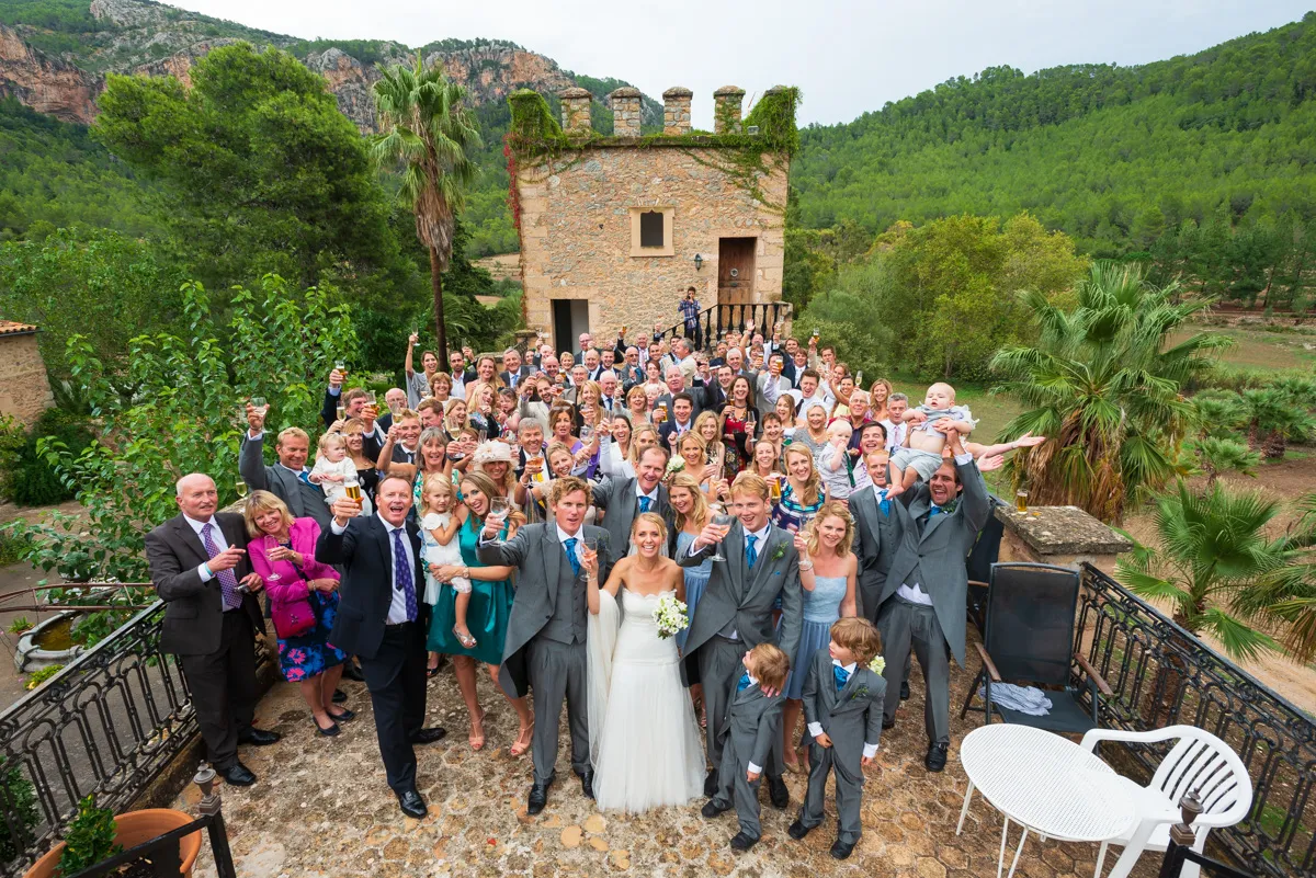 Large group of wedding guests posing with the bride and groom outdoors in front of a stone building with greenery in the background.