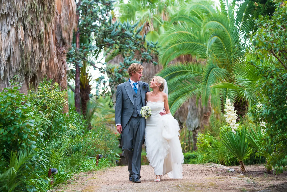 Bride in white dress and groom in gray suit walking arm in arm along garden path surrounded by lush greenery and palm trees.