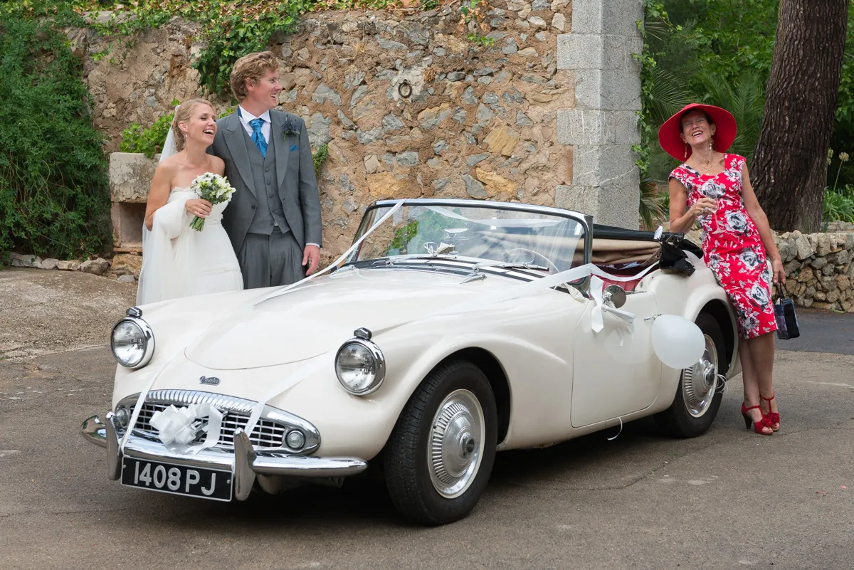 Smiling bride and groom standing beside a white vintage convertible decorated with ribbons and balloons, with a woman in a red floral dress and wide-brimmed hat holding a glass on the right.