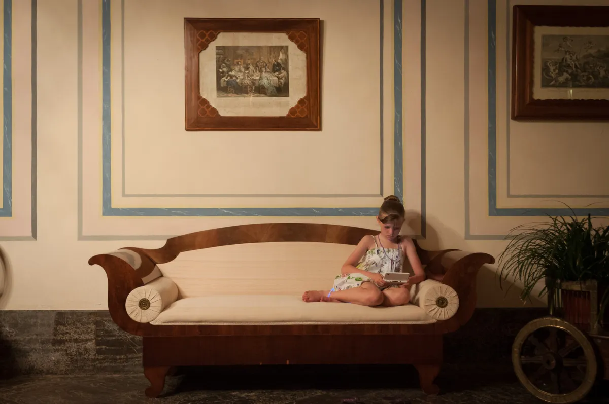 Young girl in a floral dress sitting cross-legged on a vintage wooden sofa playing a handheld gaming device in a room with framed paintings and potted plant.