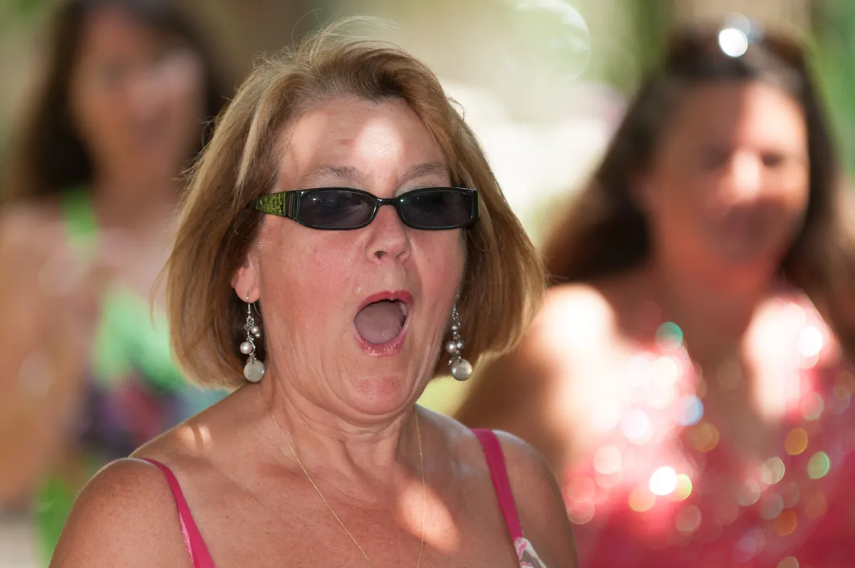 Woman wearing sunglasses and dangling earrings with mouth open in a surprised or excited expression, with two blurred women in the background.