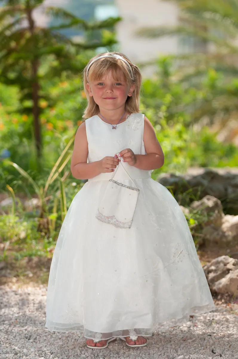 Young girl in a white dress and headband holding a small white purse, standing outdoors on gravel with greenery in the background.