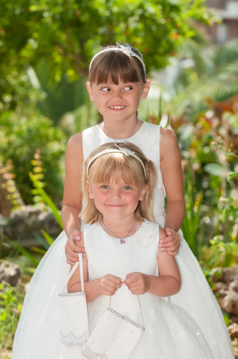 Two young girls in white dresses and headbands, standing outdoors with greenery in the background, smiling and holding small white purses.