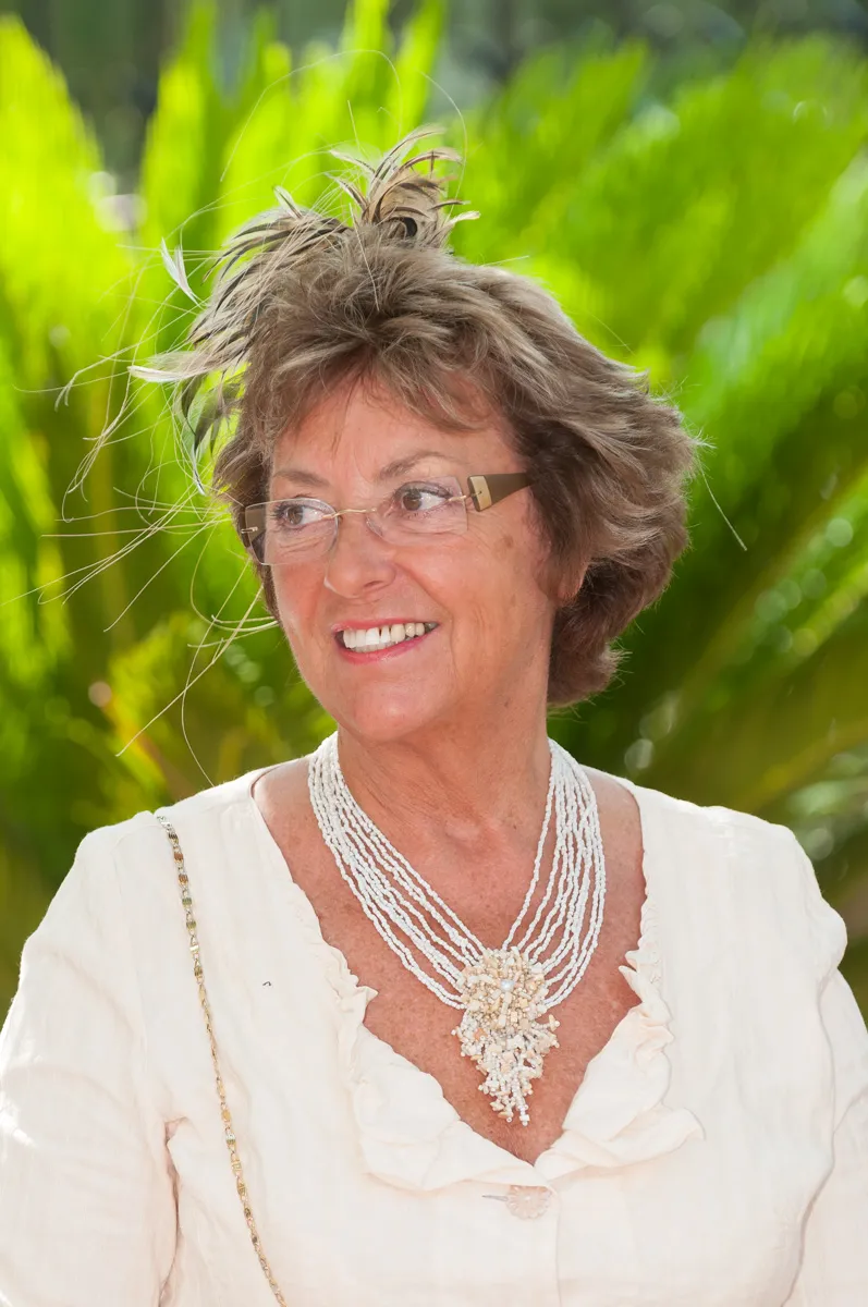 Smiling older woman with glasses, short tousled hair, wearing a white blouse and ornate beaded necklace, standing outdoors with green foliage background.