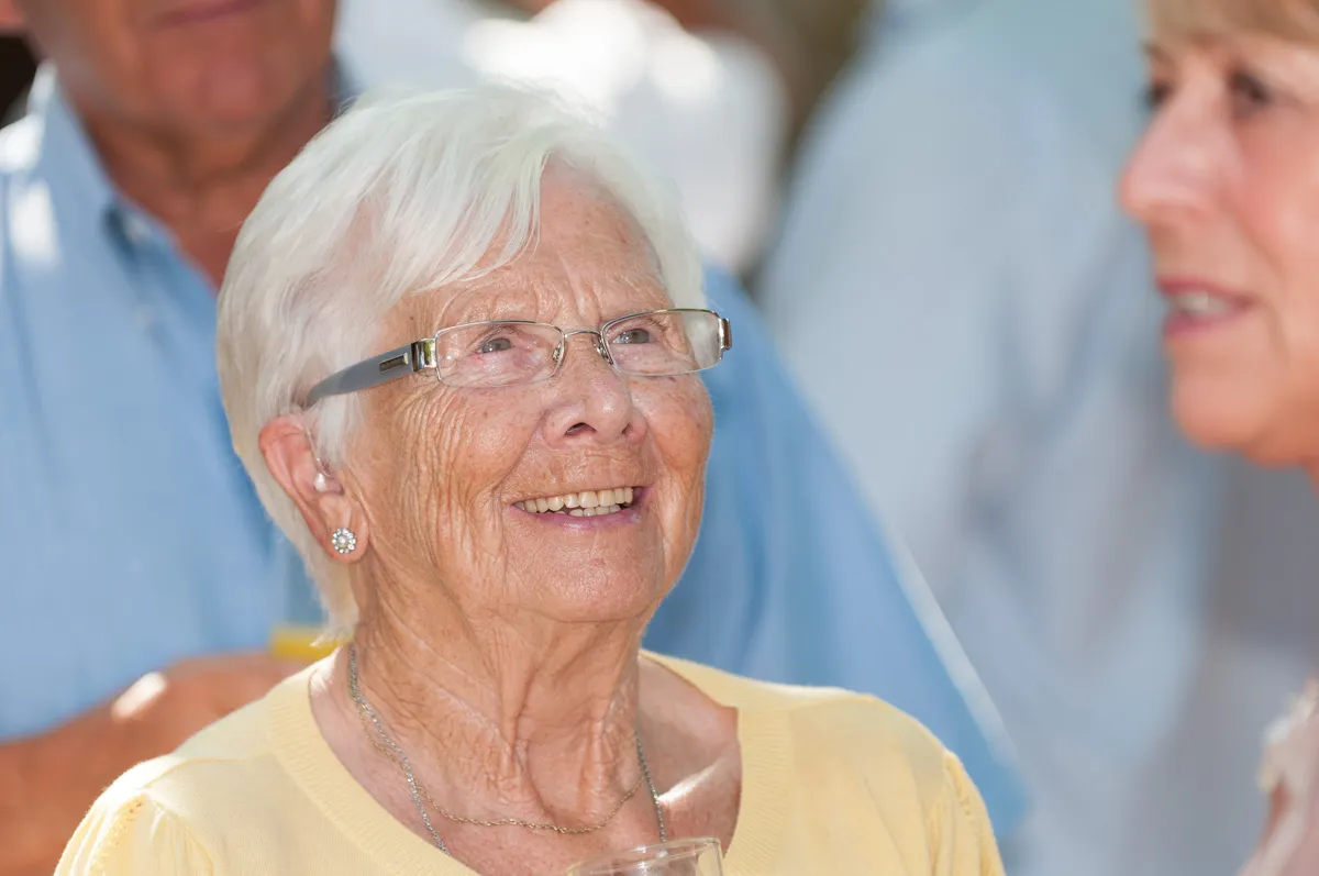 Smiling elderly woman with white hair and glasses holding a glass, wearing a yellow top and silver necklace, interacting with another person.