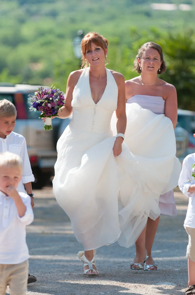 Bride in a white gown holding a purple flower bouquet walking outdoors with a bridesmaid and two young boys nearby.