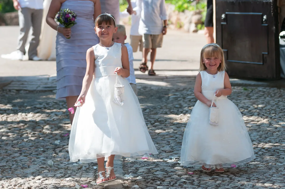 Two young flower girls in white dresses smiling and walking on a cobblestone path, each holding a small white basket with flower petals.