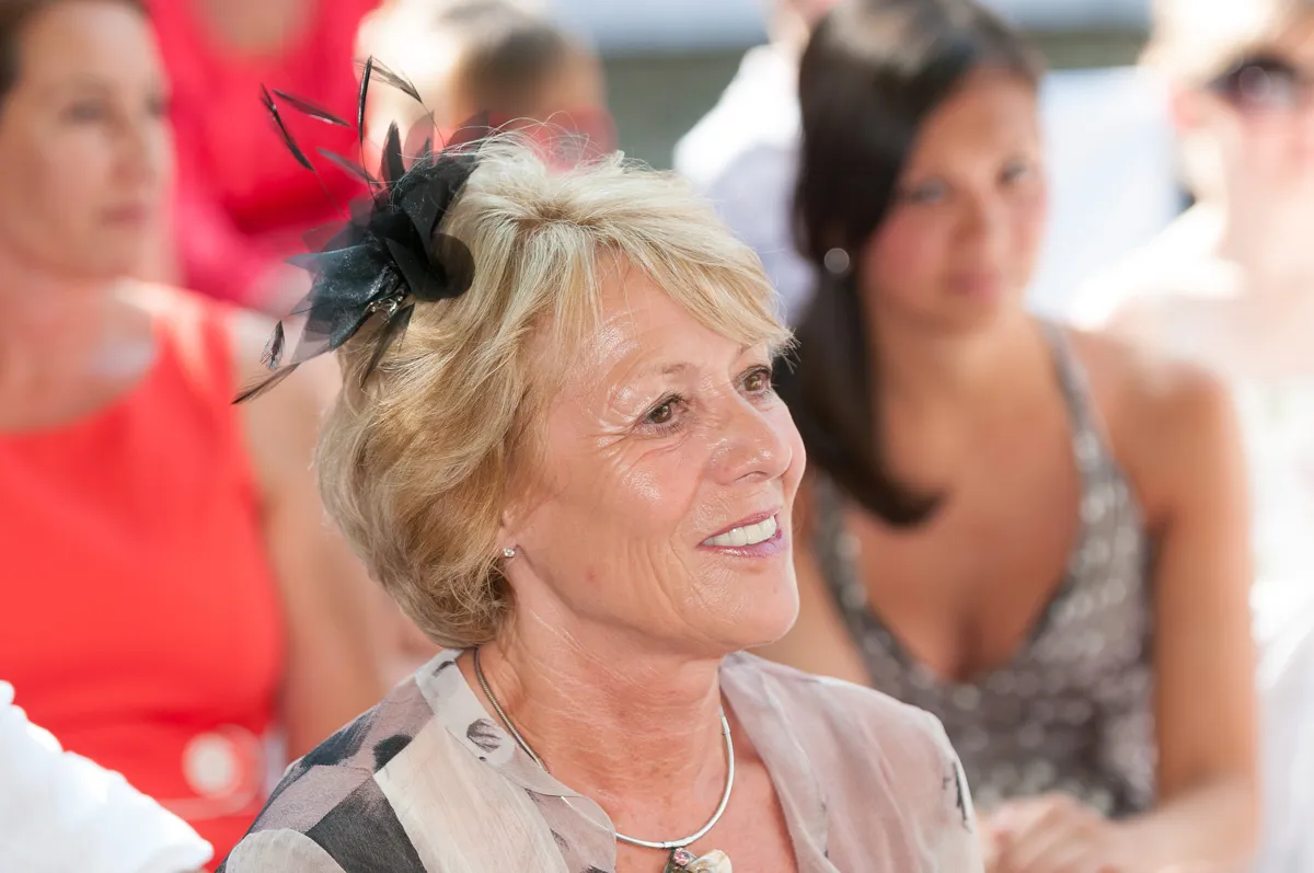 Smiling older woman wearing a black fascinator and necklace at a social event with blurred people in the background.