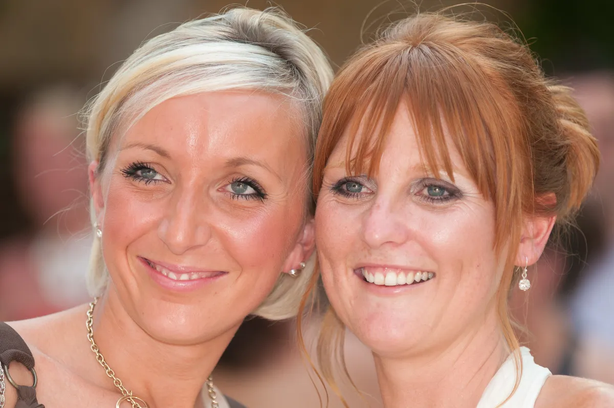 Close-up of two smiling women with light makeup, one with short blonde hair and the other with red hair tied back.