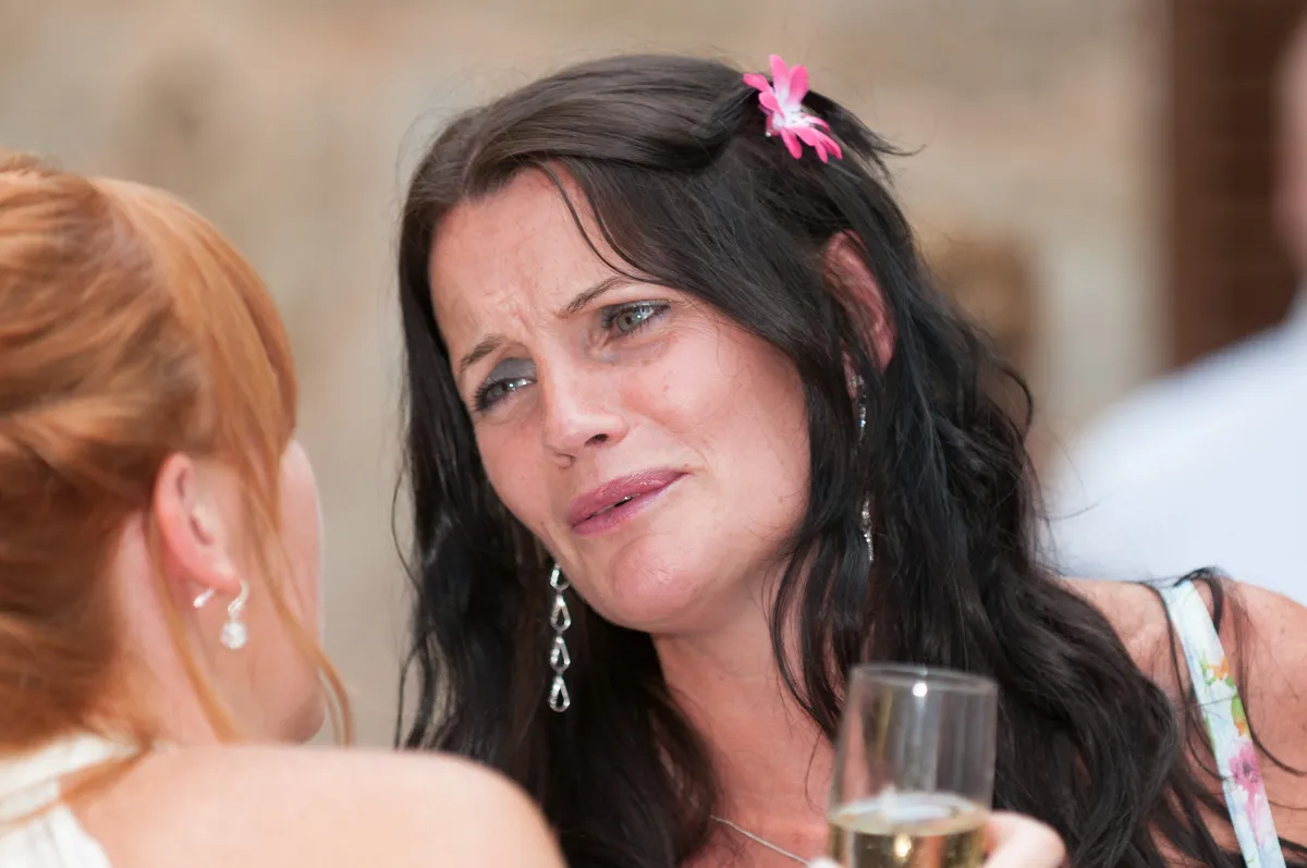 Close-up of a woman with long dark hair and a pink flower in her hair, holding a glass and speaking with another woman.