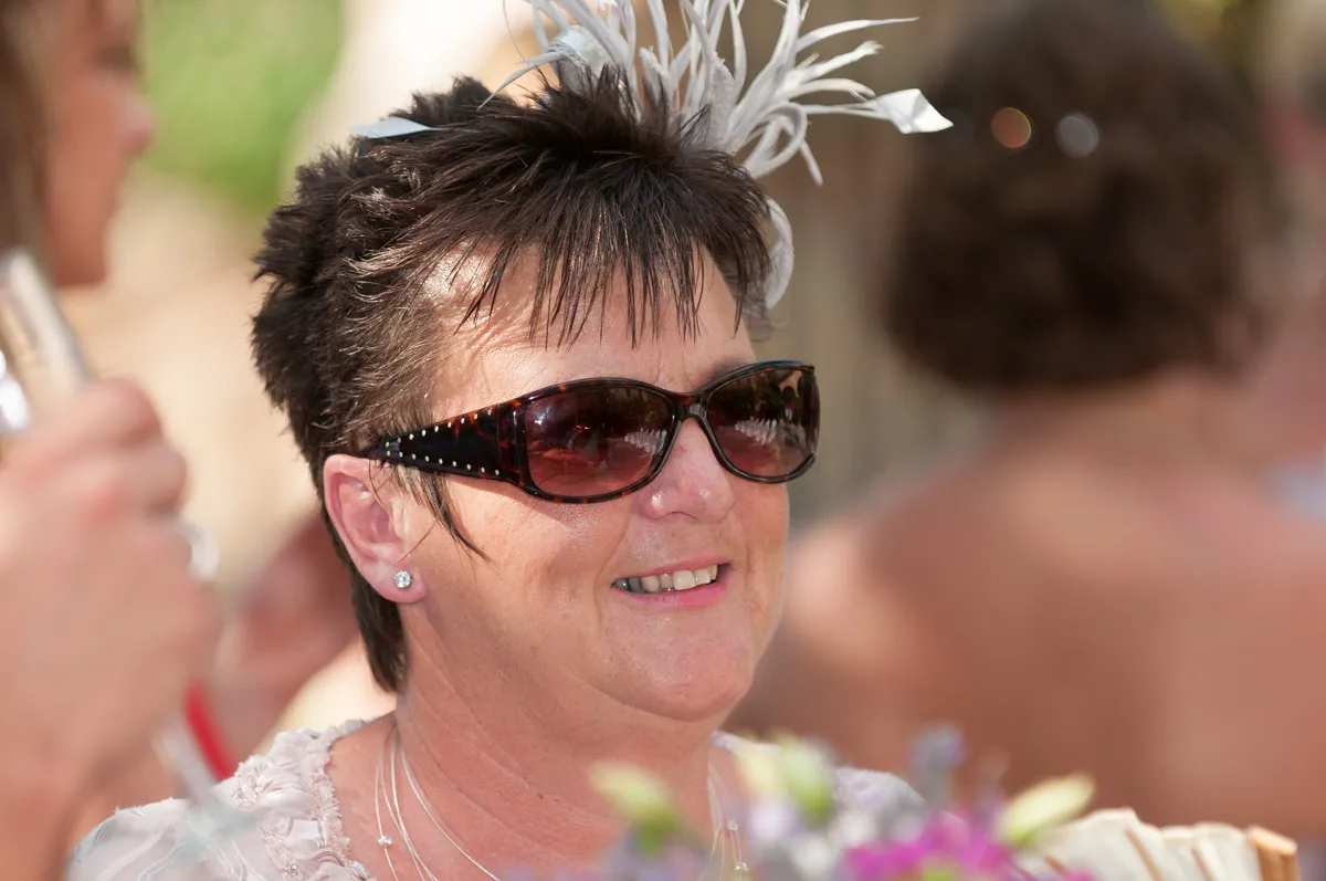 Smiling woman wearing dark sunglasses and a white feathered fascinator, with a floral bouquet in the foreground.