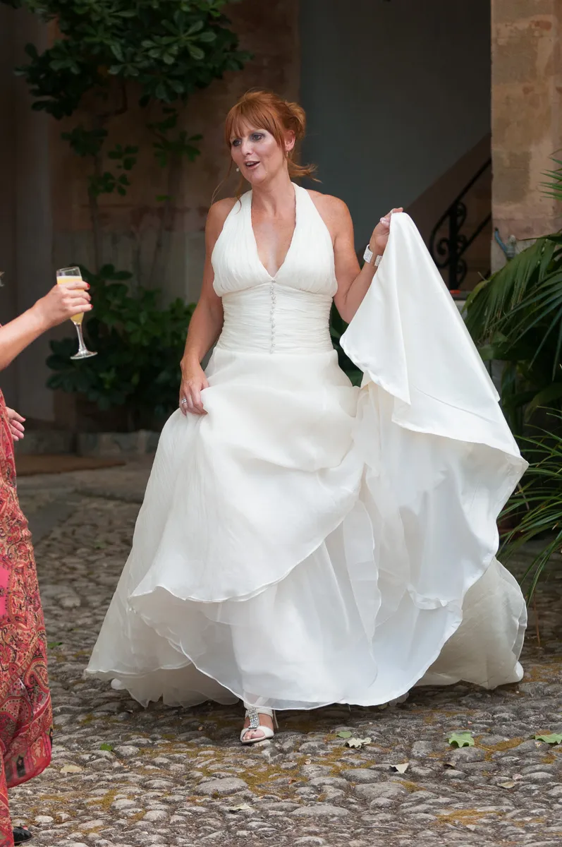Red-haired bride in a sleeveless white wedding gown lifting her skirt while standing on a cobblestone path outdoors.