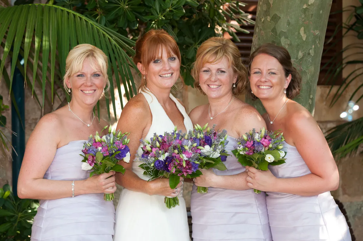 Bride in white dress and three bridesmaids in light lavender dresses holding colorful purple and white flower bouquets, smiling outdoors with green foliage background.