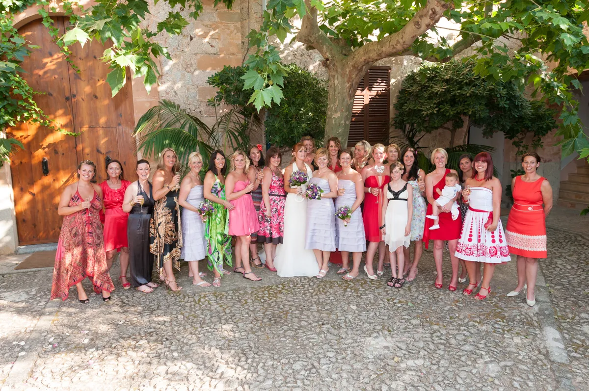 Group of women and a child in colorful dresses posing outdoors on a stone pathway under green leafy trees.