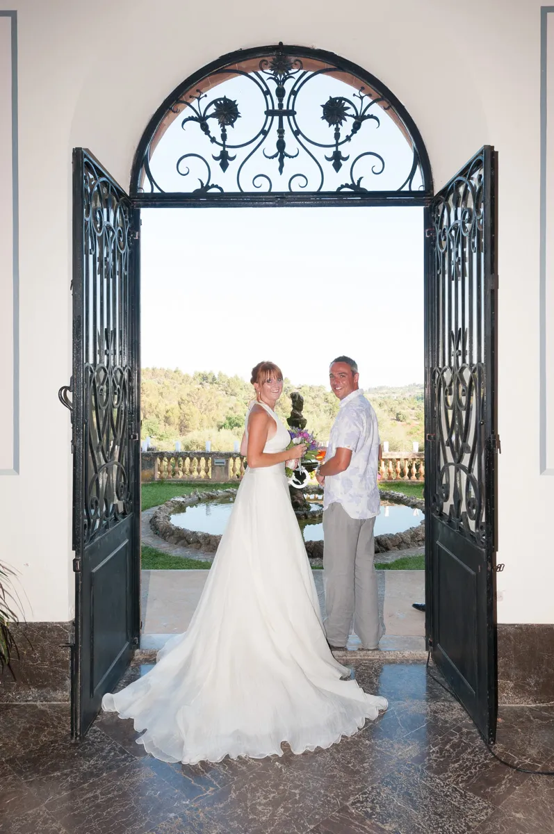 Bride in a white wedding gown and a man standing in front of open ornate black iron doors looking back, with a garden and fountain in the background.