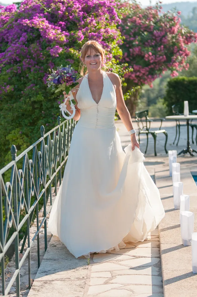 Smiling bride in a white halter wedding dress holding a bouquet, walking along an outdoor stone path with purple flowering bushes in the background.