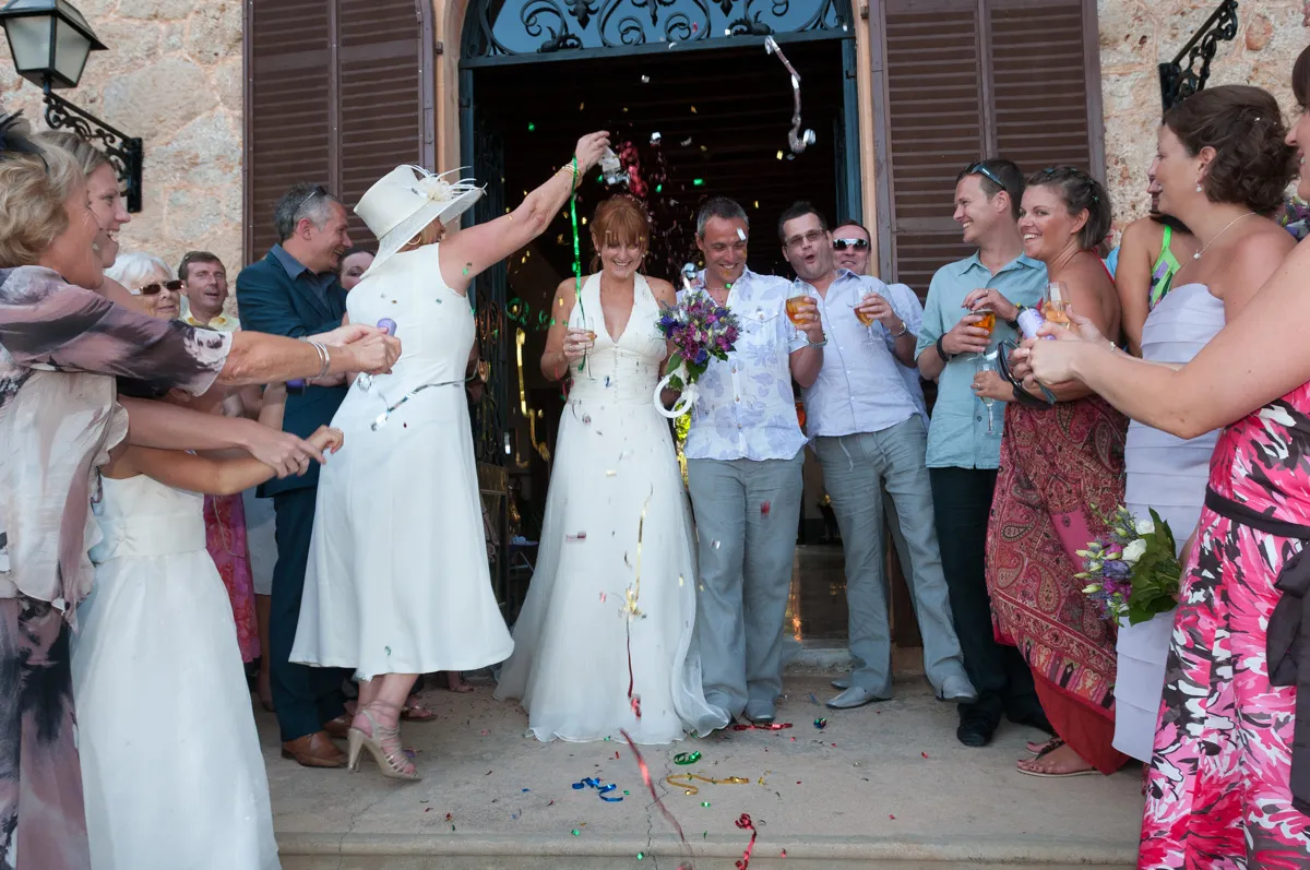 Bride and groom smiling and holding drinks while guests celebrate with confetti outside a building.