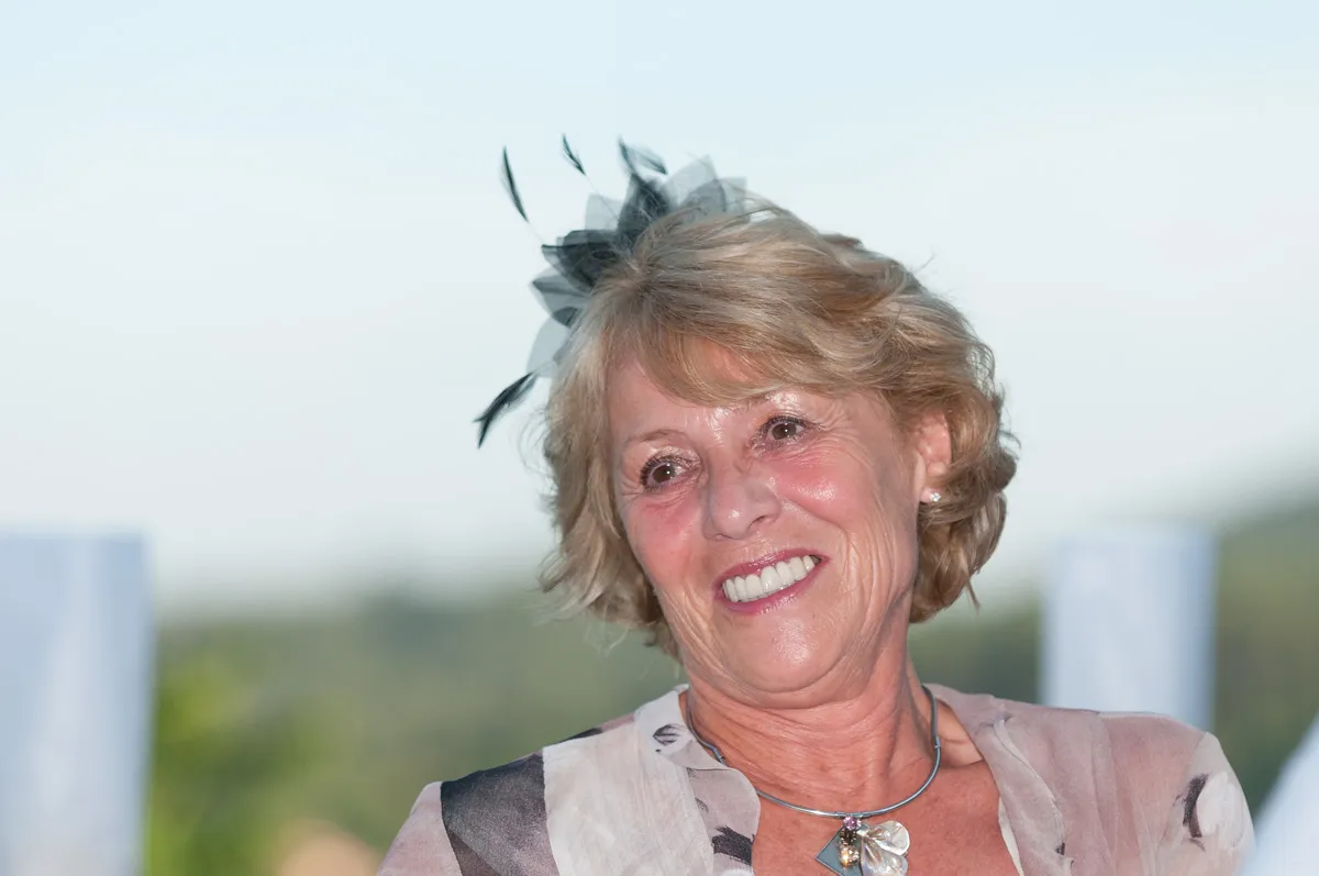 Smiling older woman with short blonde hair wearing a black feathered hair accessory and a silver necklace outdoors.