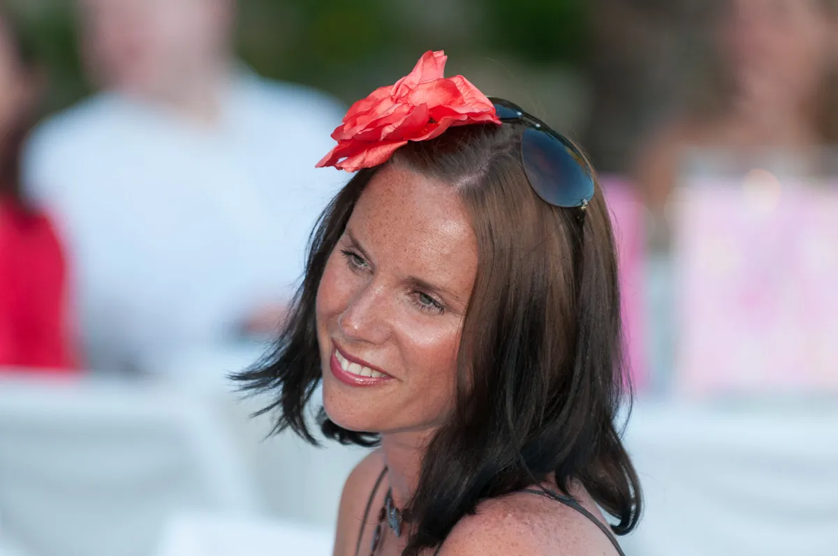 Smiling woman with shoulder-length brown hair, wearing sunglasses on her head and a large red flower in her hair.
