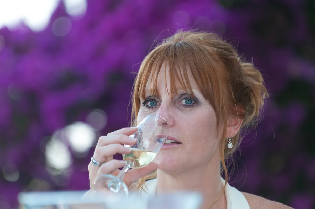 Close-up of a woman with red hair and blue eyes sipping white wine from a glass with a blurred purple floral background.
