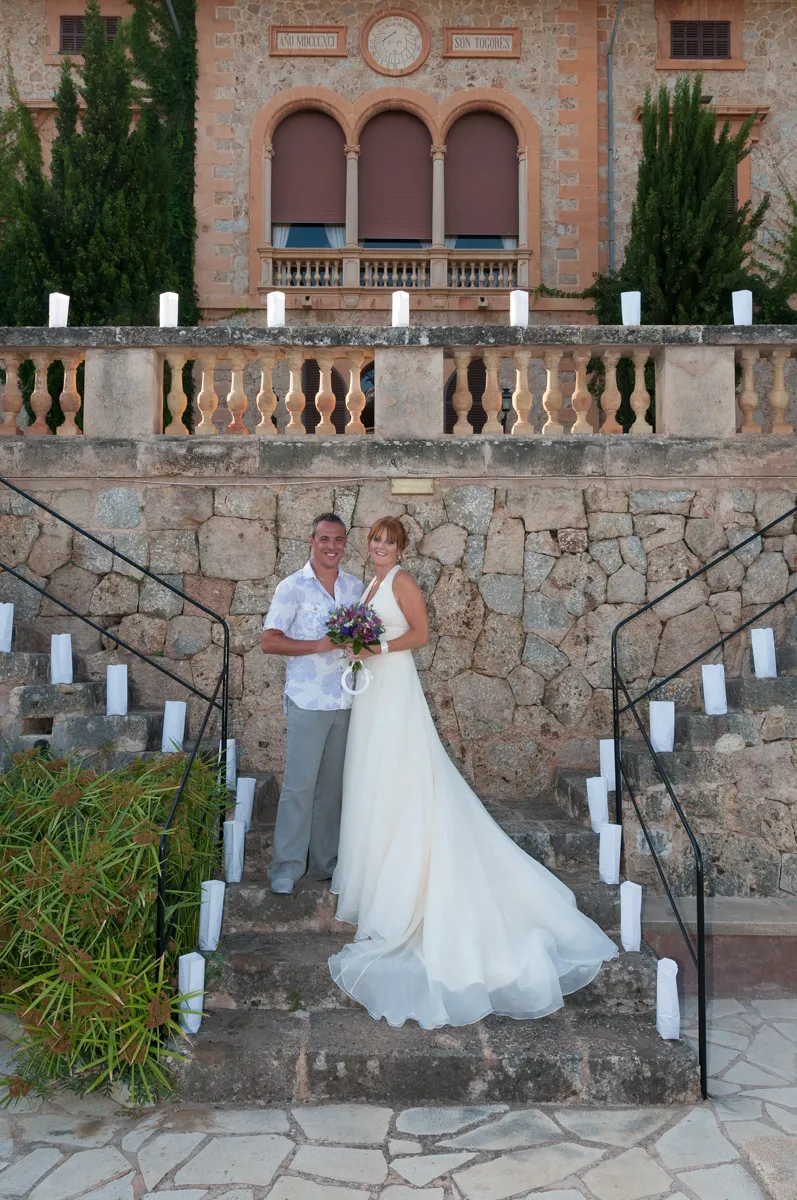 Bride in white wedding dress and groom in casual shirt and pants standing on stone steps in front of a rustic stone building.