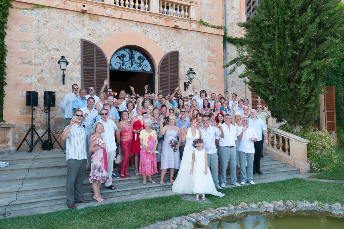 Large group of people, including a bride in a wedding dress holding a bouquet and a girl in a white dress, standing on steps outside a stone building with arched doors, raising glasses in celebration.