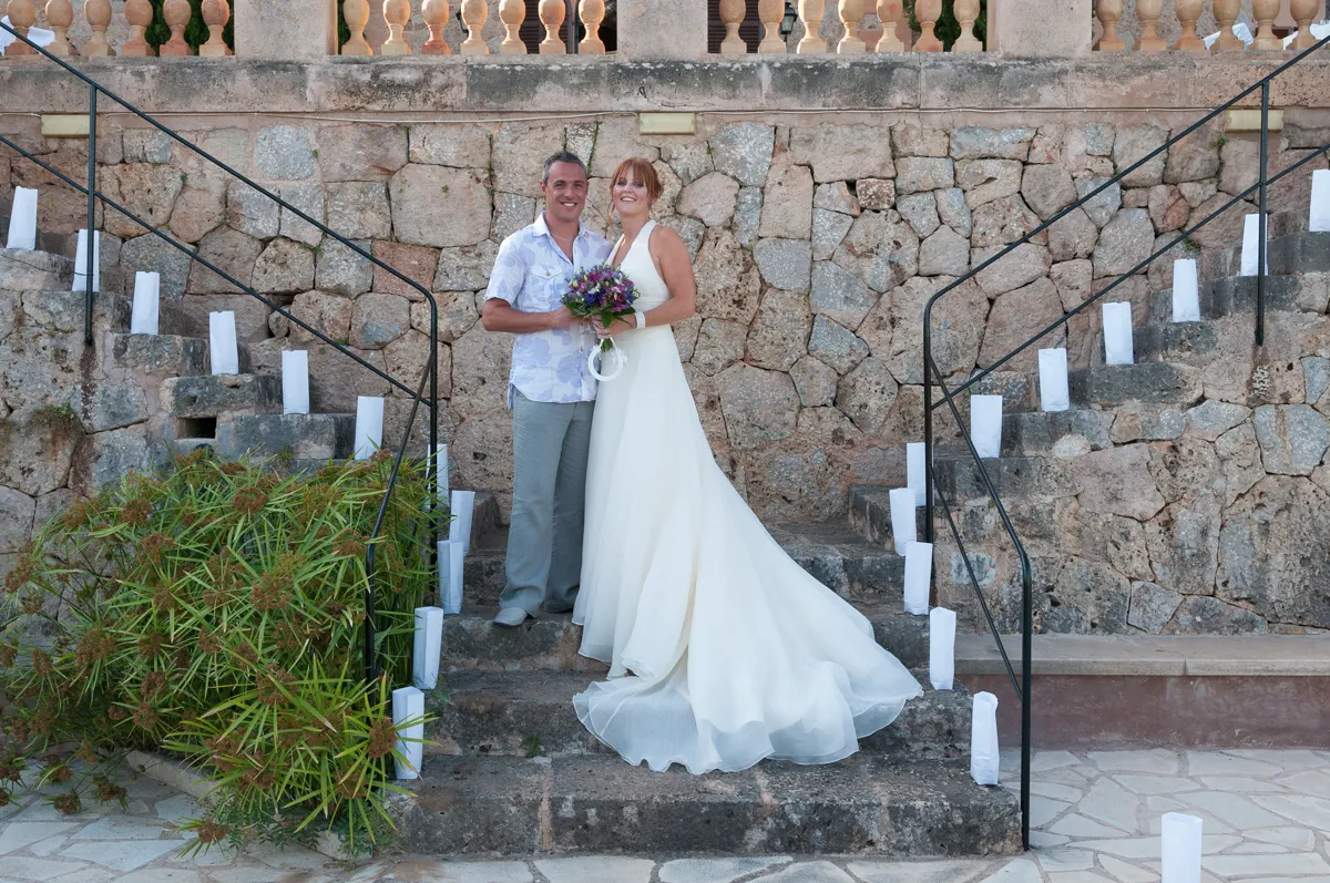 Bride in a flowing white gown holding a purple bouquet stands next to a smiling groom in a light shirt and gray pants on stone steps with white candleholders and a stone wall backdrop.