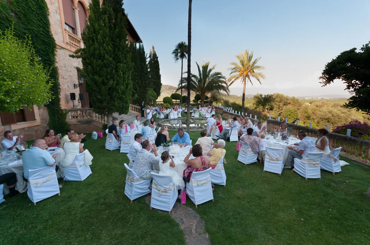 Outdoor wedding reception with guests seated at round tables on a lawn beside a stone building and palm trees.