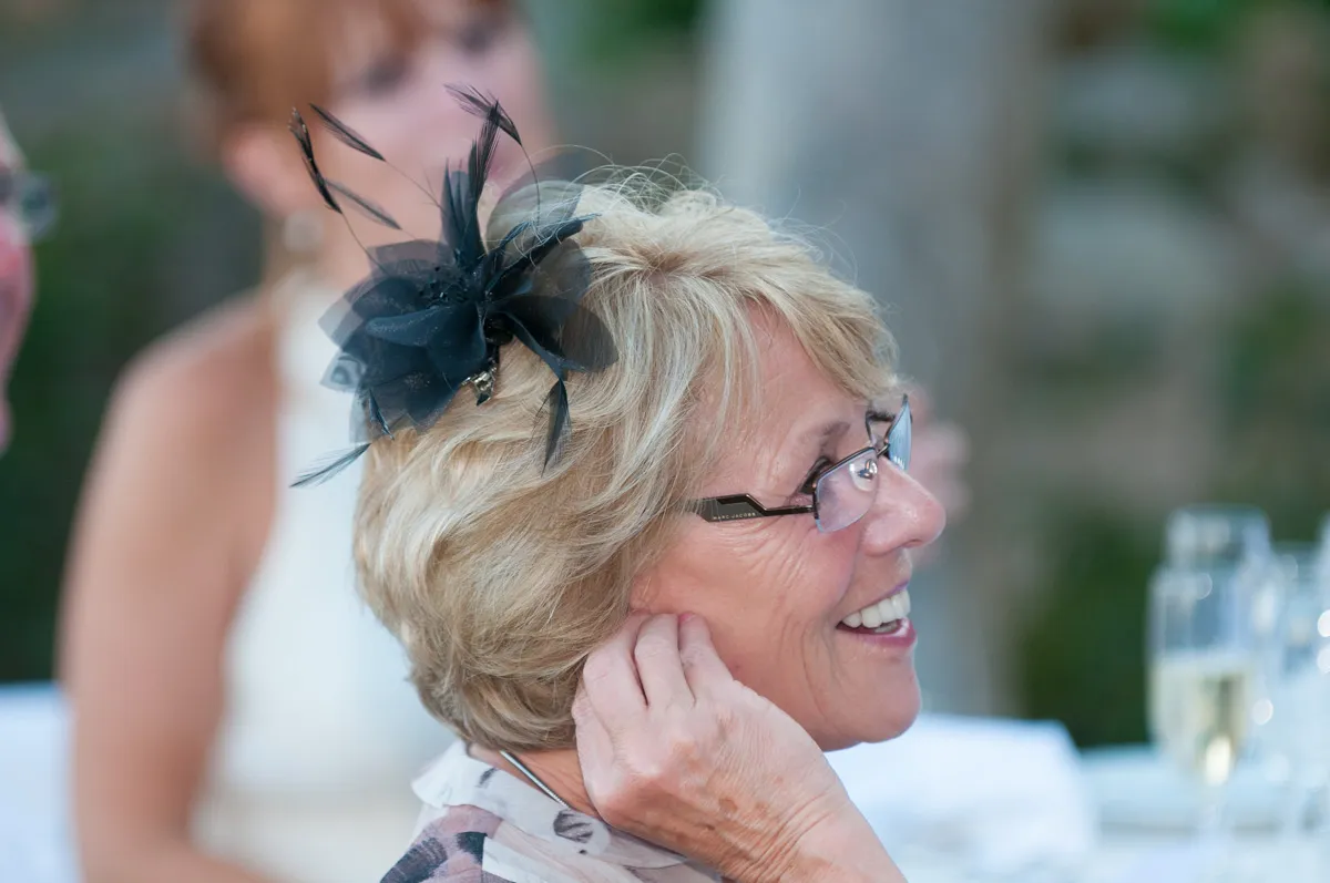 Smiling older woman with blond hair wearing glasses and a black feathered fascinator, touching her ear at a social event.
