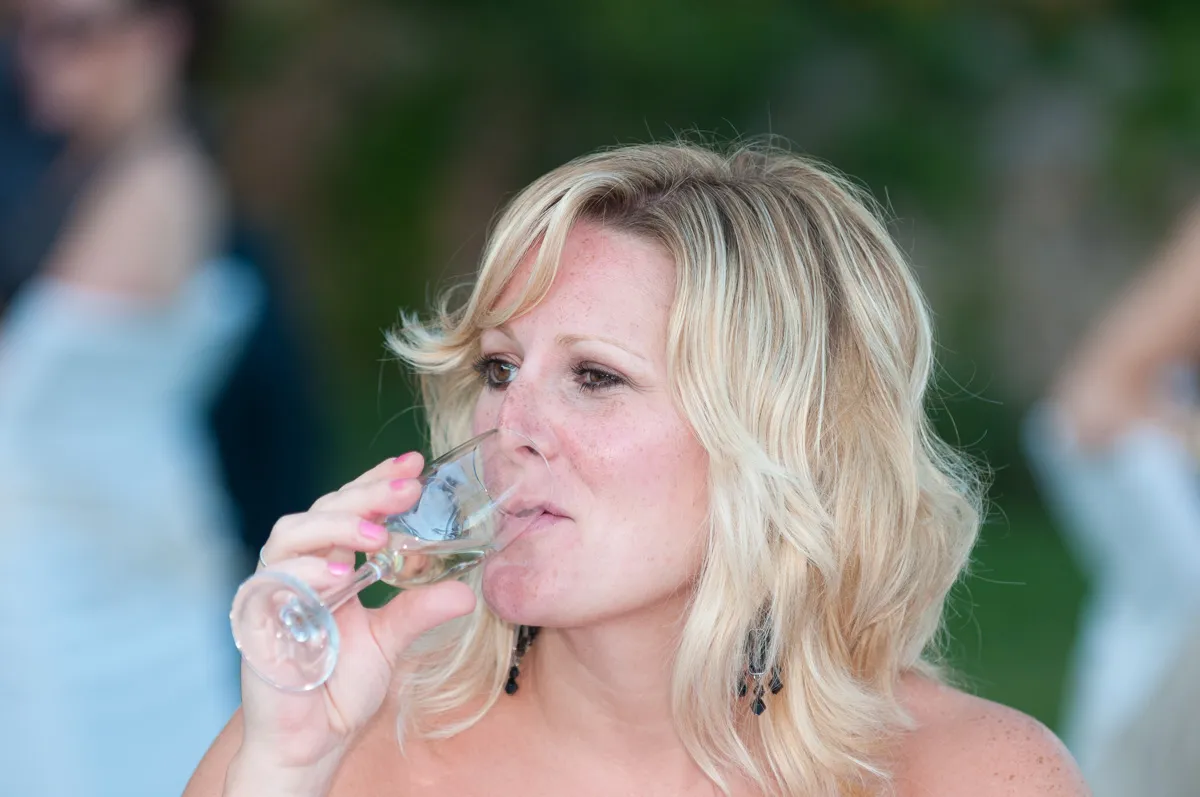Blonde woman with wavy hair and black earrings sipping a drink from a wine glass outdoors.