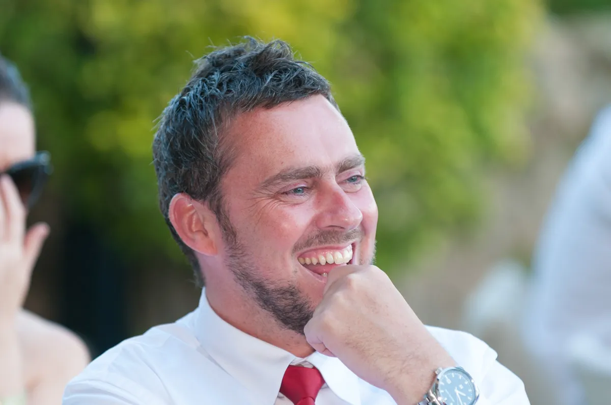 Smiling man in white shirt and red tie resting his chin on his hand with blurred greenery background.