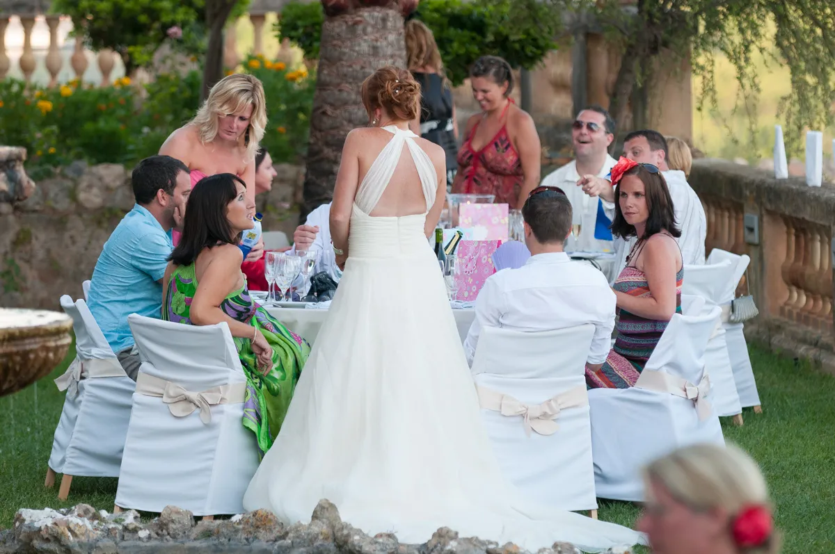 Bride in white gown standing and talking to guests seated at an outdoor table with white-covered chairs during a garden wedding reception.