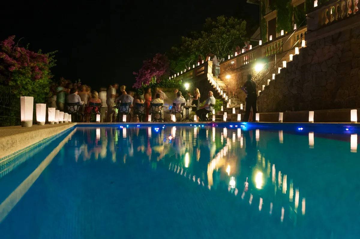 Nighttime gathering by a pool with people seated and standing near lit lanterns along the pool edge and stairs.