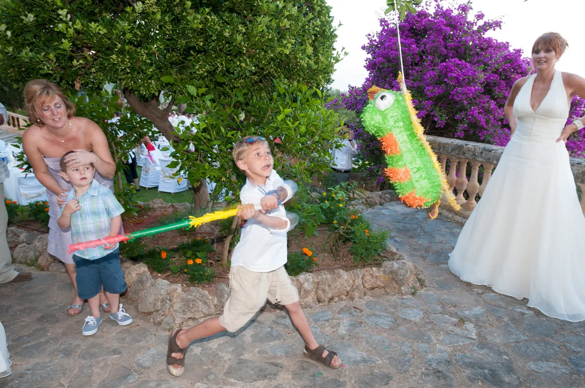 A young boy swings a colorful stick at a green and orange piñata shaped like a bird while a woman in a white dress and a woman with a young child watch nearby.