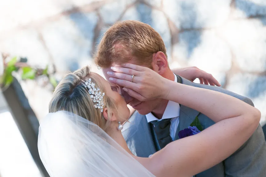 Bride with veil and decorative hairpiece gently covering groom's eyes as they embrace closely.