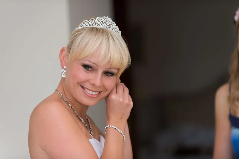 Smiling blonde bride wearing a tiara and white strapless dress, adjusting an earring.