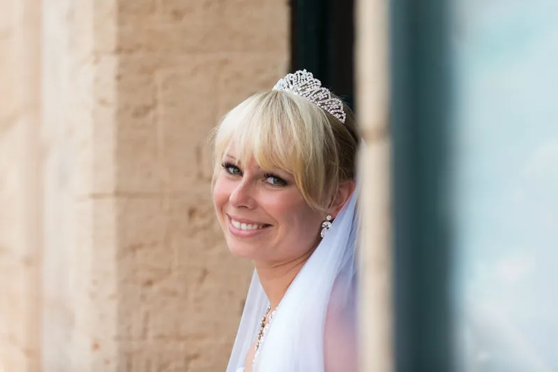 Smiling bride with blonde hair wearing a tiara and veil, leaning against a stone wall.