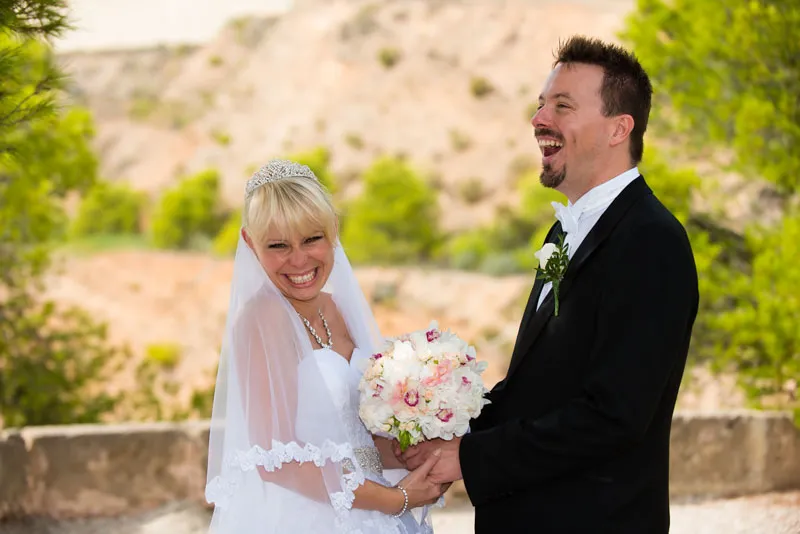 Bride and groom smiling and holding hands outdoors with the bride holding a bouquet of white and pink flowers.