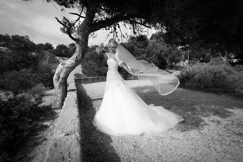 Bride in a wedding dress and veil smiling outdoors under a tree beside a stone wall.