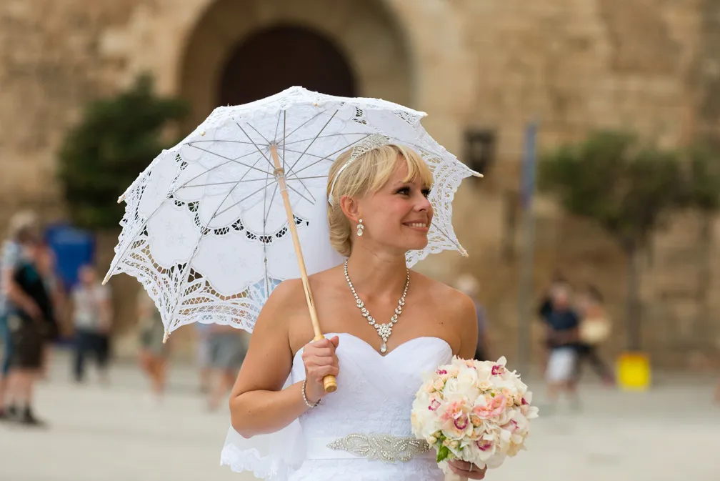 Smiling bride wearing white dress and jewelry, holding a lace parasol and a bouquet of flowers outdoors.